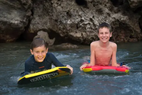 Two boys smiling on bodyboards near rocky coastline